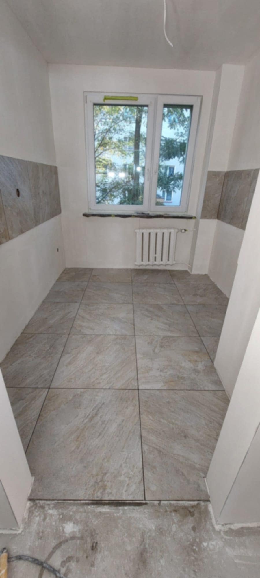 Wykończona podłoga z płytek w jasnym odcieniu, fragment ściany z dekoracyjnymi panelami, okno z widokiem na zieleń. Grzejnik pod oknem.