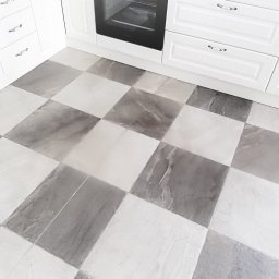 Wykończenia Wnętrz Marian Ryba - Szachownica z płytek podłogowych w kuchni z białymi szafkami i piekarnikiem w tle. Płytki w odcieniach szarości i bieli tworzą wzór. Ujęcie z góry, skupione na podłodze.