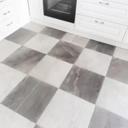 Szachownica z płytek podłogowych w kuchni z białymi szafkami i piekarnikiem w tle. Płytki w odcieniach szarości i bieli tworzą wzór. Ujęcie z góry, skupione na podłodze.