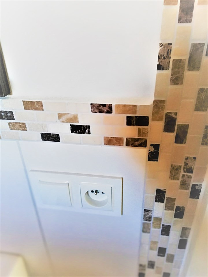 Wykończenie ściany mozaiką z elementami imitującymi marmur, gniazdko elektryczne i włącznik światła na białej ścianie. Ukończony etap prac wykończeniowych.
