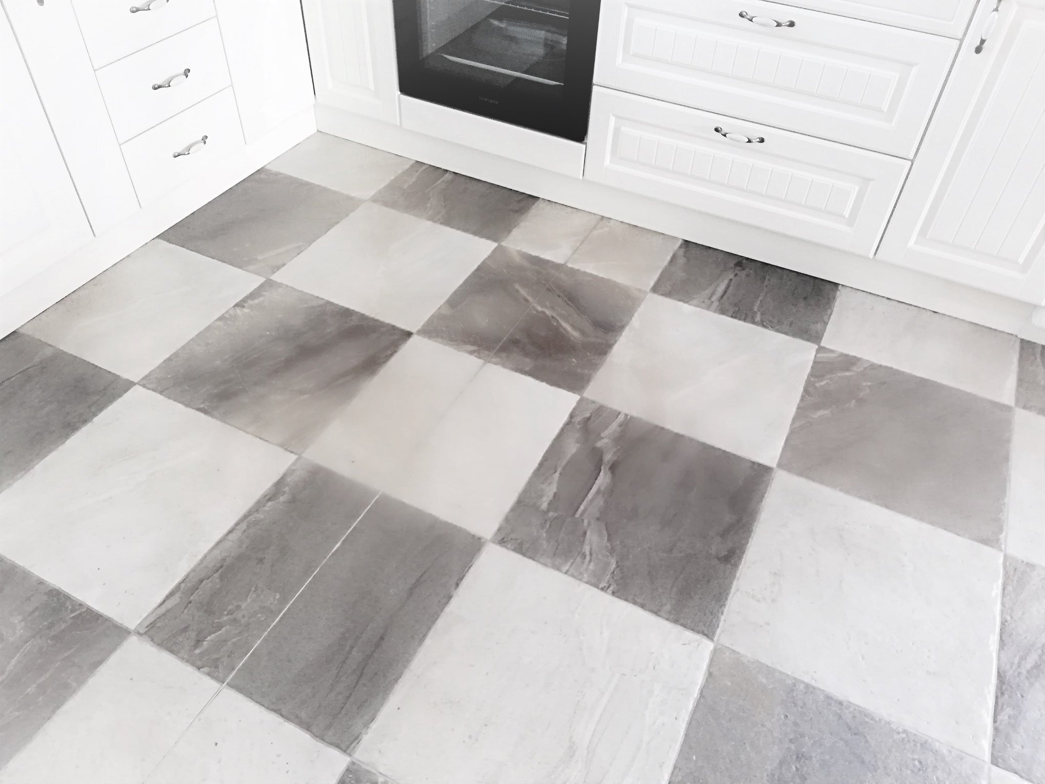 Szachownica z płytek podłogowych w kuchni z białymi szafkami i piekarnikiem w tle. Płytki w odcieniach szarości i bieli tworzą wzór. Ujęcie z góry, skupione na podłodze.