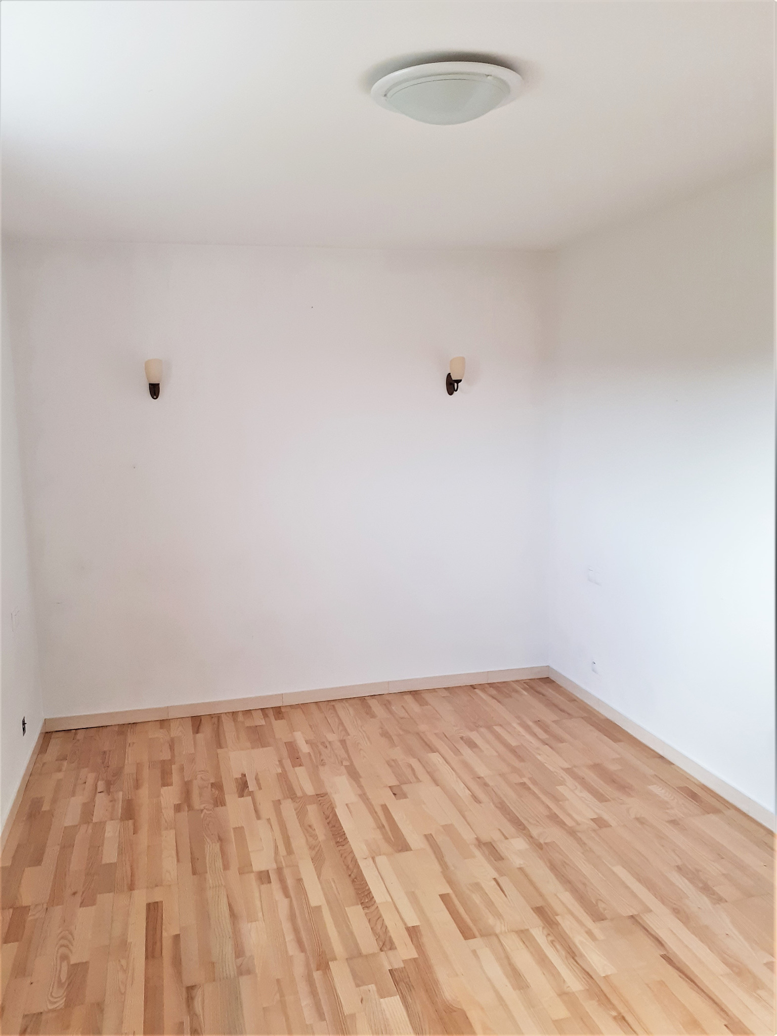 Pusty, jasny pokój po remoncie z parkietem, białymi ścianami i dwoma kinkietami w stylu retro. Widok z góry na odnowione wnętrze.