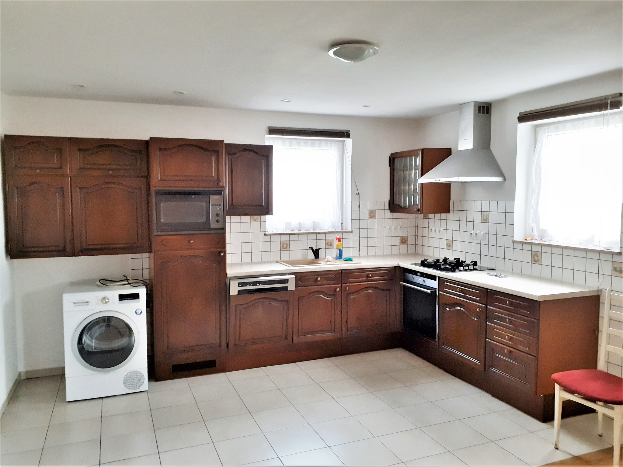 Jasna kuchnia w stylu retro z ciemnymi, drewnianymi szafkami, białymi płytkami na ścianach i podłodze. Biała pralka wolnostojąca obok zabudowy kuchennej.