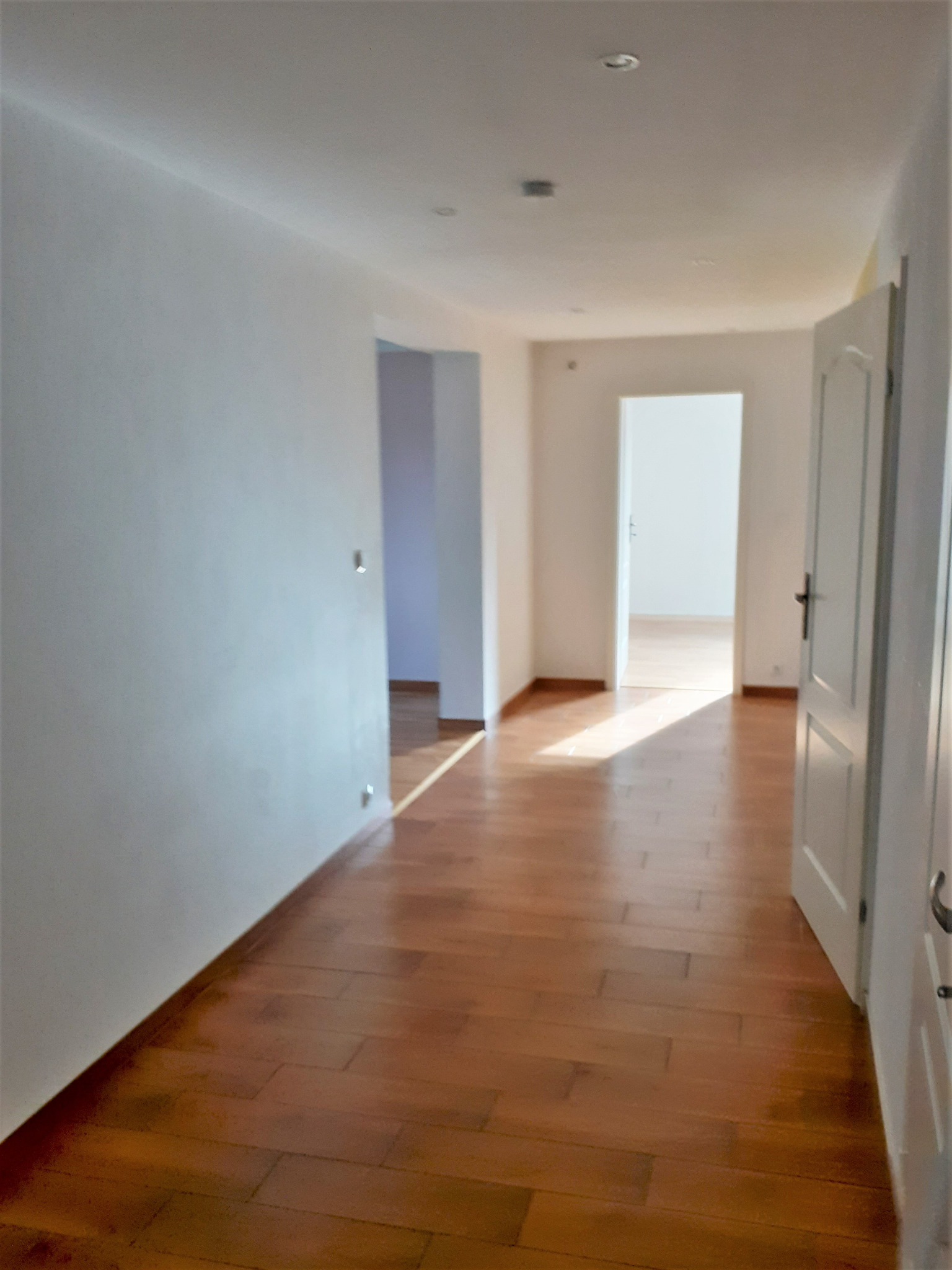 Wykończony korytarz z parkietem i świeżo pomalowanymi ścianami na biało. Otwarta przestrzeń z widokiem na dalsze pomieszczenia. Drzwi wewnętrzne otwarte.