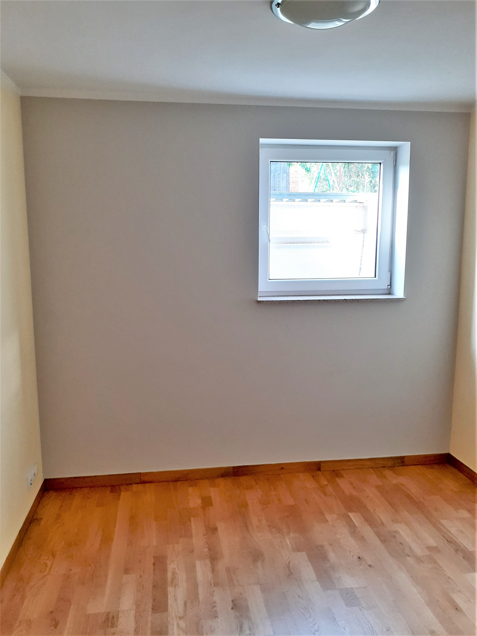 Jasne wnętrze pokoju po remoncie: drewniana podłoga, beżowe ściany, białe okno z widokiem na zieleń. Minimalistyczne wykończenie, gotowe do aranżacji.