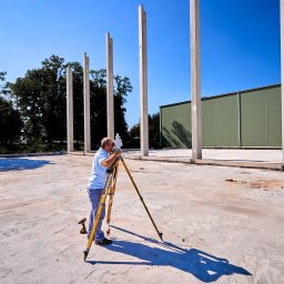 USŁUGI GEODEZYJNE - MARIUSZ FIT - Pracownik z niwelatorem na budowie, mierzący teren pod budowę hali. Widoczne betonowe słupy i zielona ściana hali w tle, na tle błękitnego nieba.