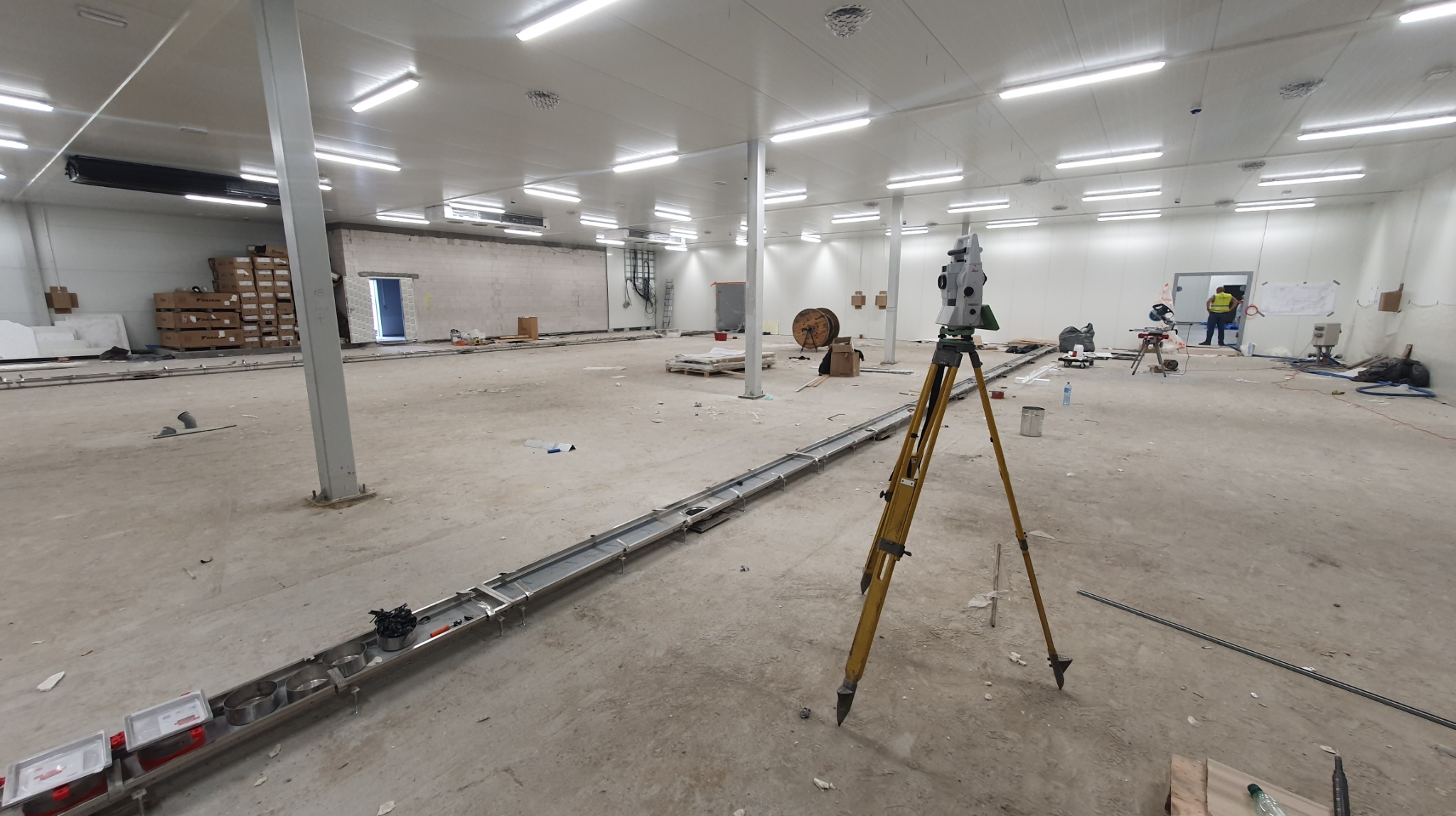 Teodolit na statywie w hali produkcyjnej, pomiary w trakcie prac instalacyjnych. Widoczne elementy konstrukcyjne i instalacje.