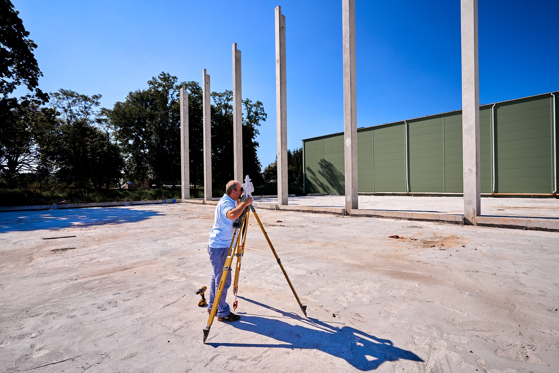 Pracownik z niwelatorem na budowie, mierzący teren pod budowę hali. Widoczne betonowe słupy i zielona ściana hali w tle, na tle błękitnego nieba.