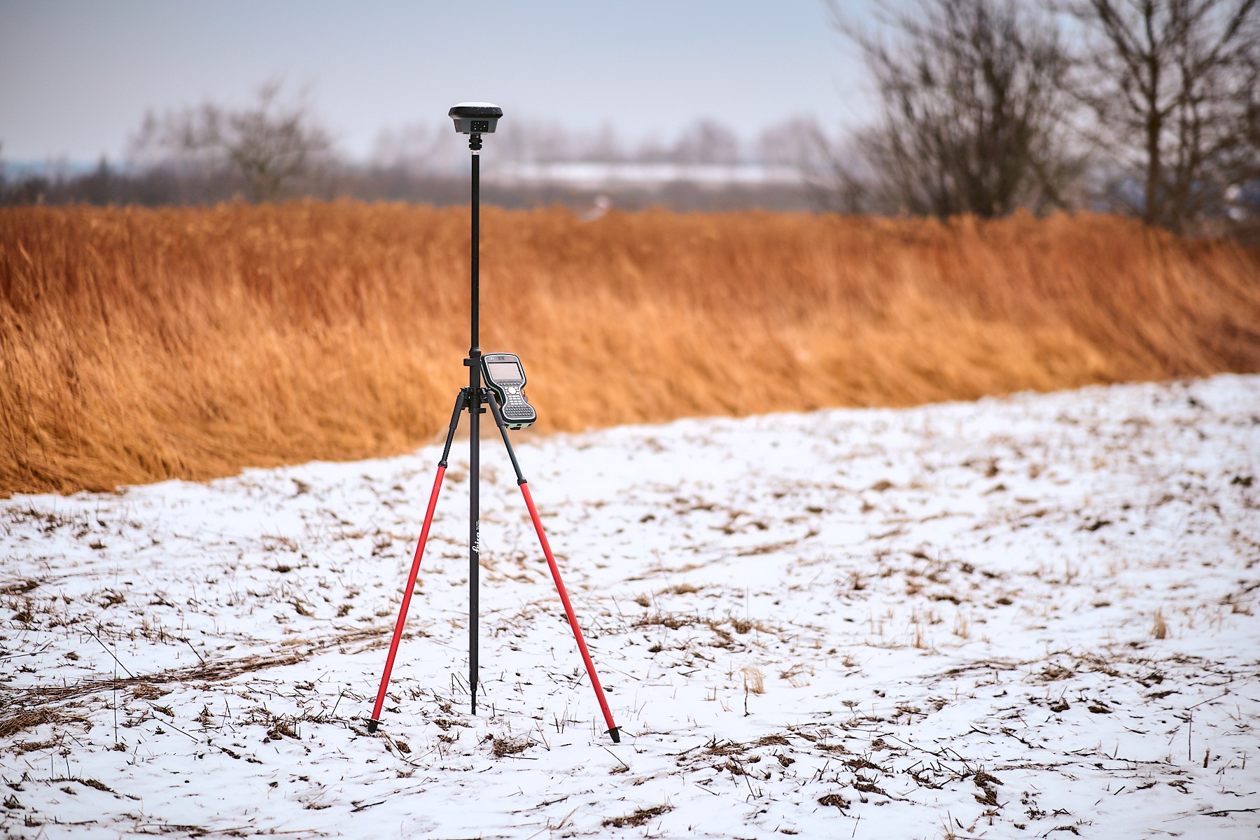 Czarny odbiornik GNSS na czerwonym statywie w zimowym krajobrazie. W tle brązowe trawy i drzewa pod pochmurnym niebem. Precyzyjny pomiar terenu.