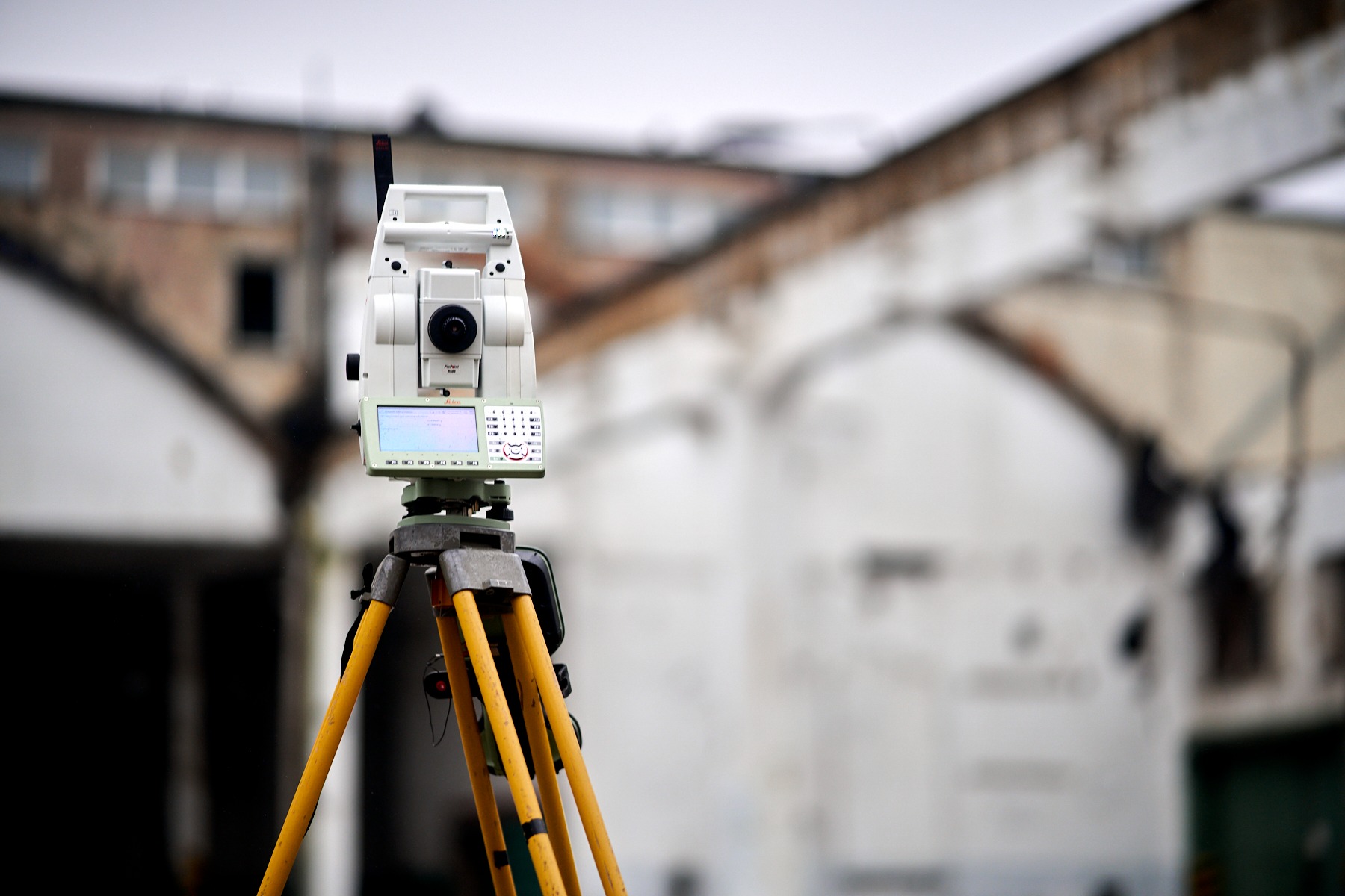 Elektroniczny tachimetr Leica na statywie z żółtymi nogami, na tle budynku z cegły. Sprzęt pomiarowy ustawiony na zewnątrz budynku.