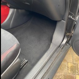 Bartłomiej karolak - Wnętrze czarnego samochodu po praniu tapicerki, widoczne czyste dywaniki i podłoga, z detalami deski rozdzielczej i siedzenia kierowcy.