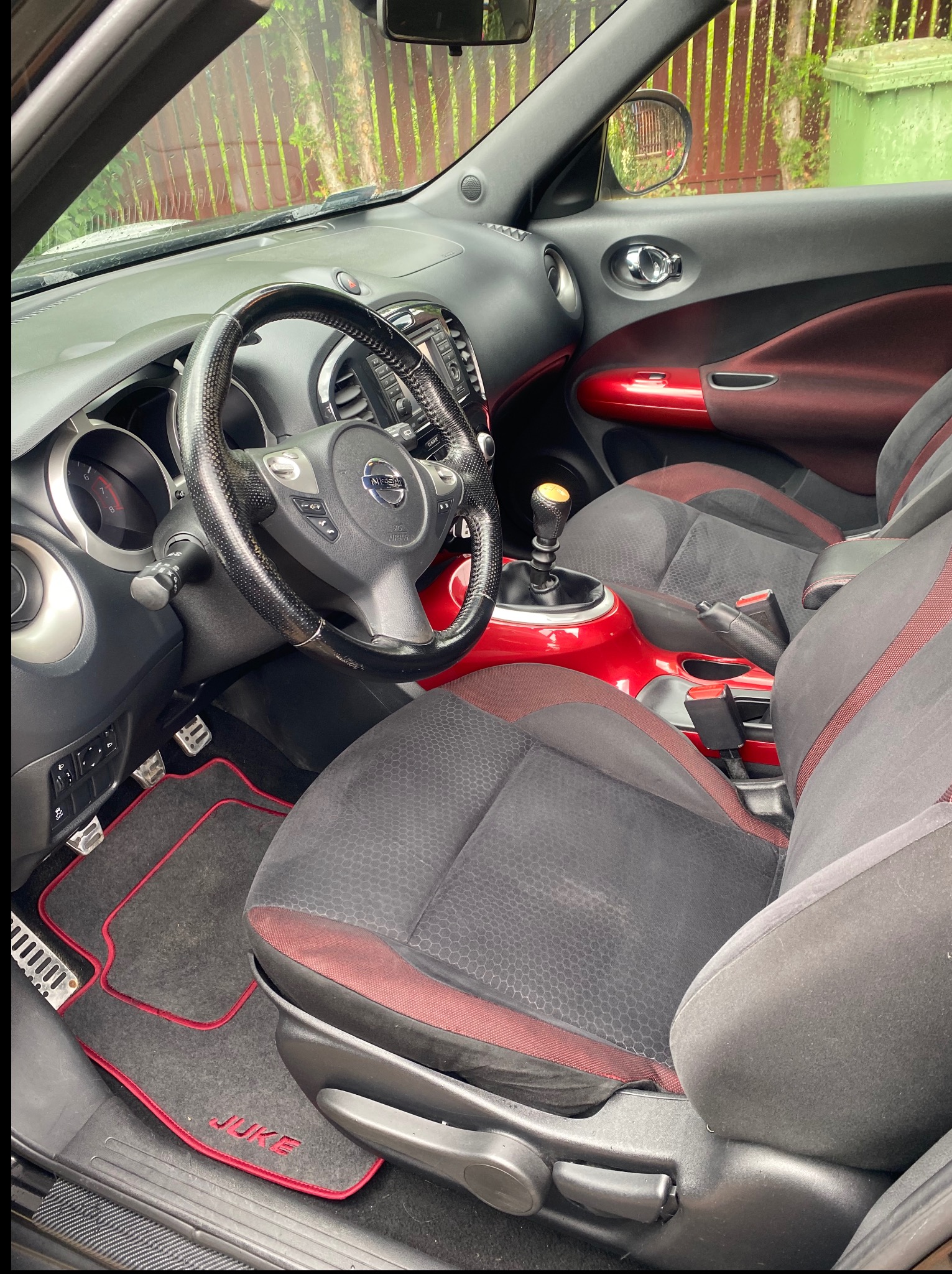 Wnętrze samochodu Nissan Juke z czarno-czerwonymi akcentami. Widoczne fotele, kierownica z karbonowym wykończeniem i dywaniki z logo Juke.