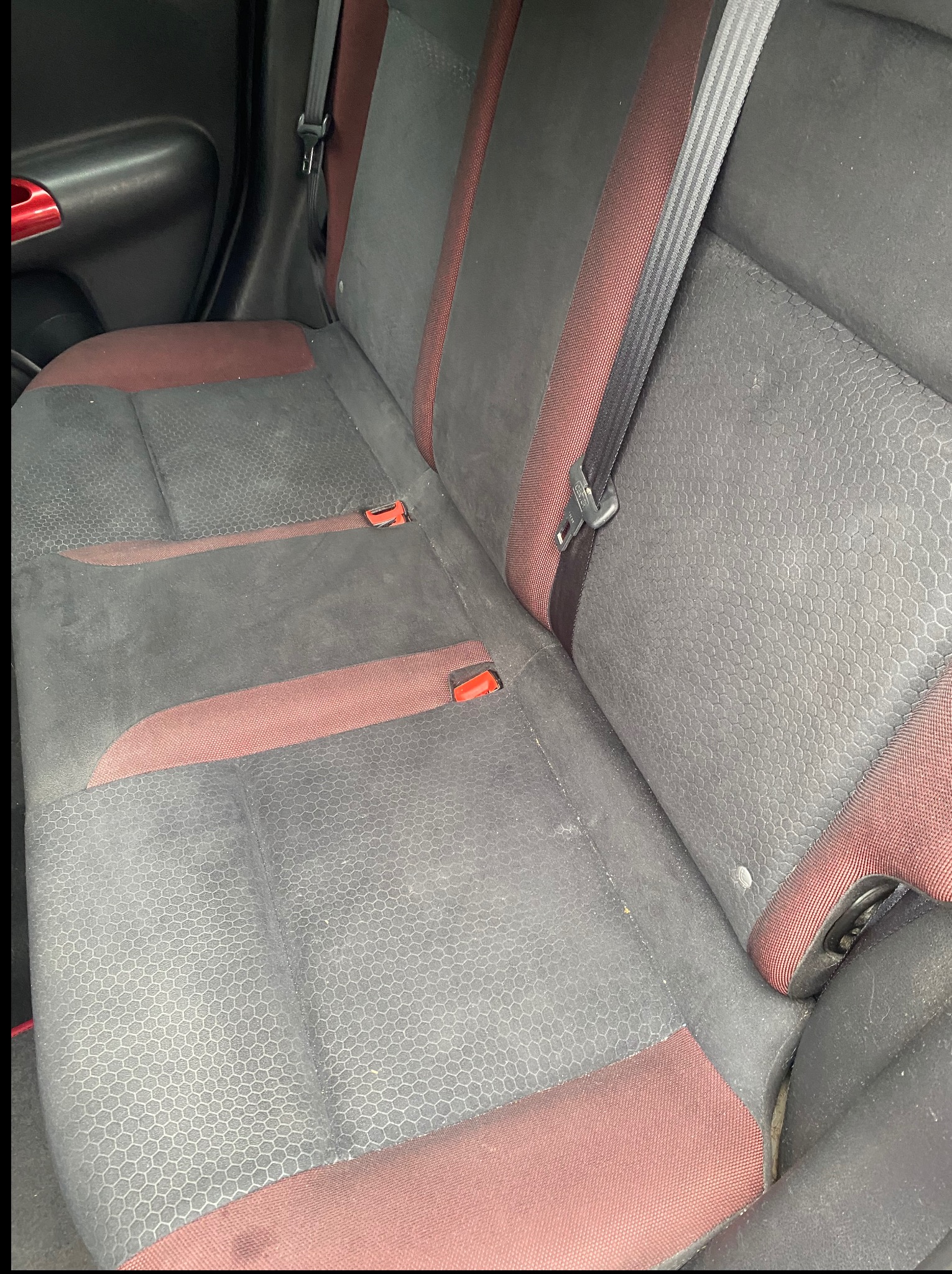 Tylna kanapa samochodu z szaro-bordową tapicerką, widoczne zabrudzenia i wzór w sześciokąty. Pas bezpieczeństwa na siedzeniu.