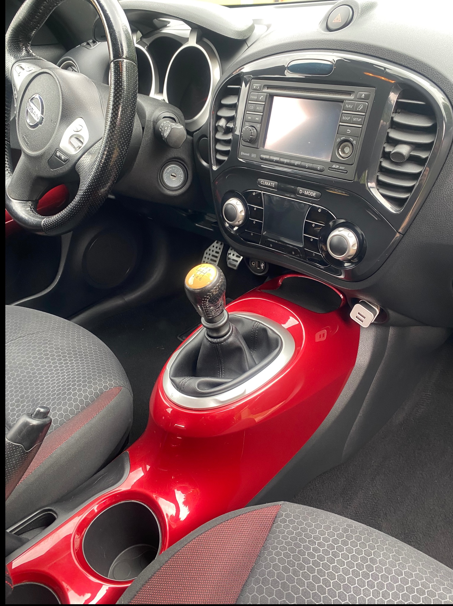 Wnętrze samochodu Nissan Juke z widocznymi detalami: kierownica, deska rozdzielcza, dźwignia zmiany biegów z żółtą gałką i czerwony panel środkowy.