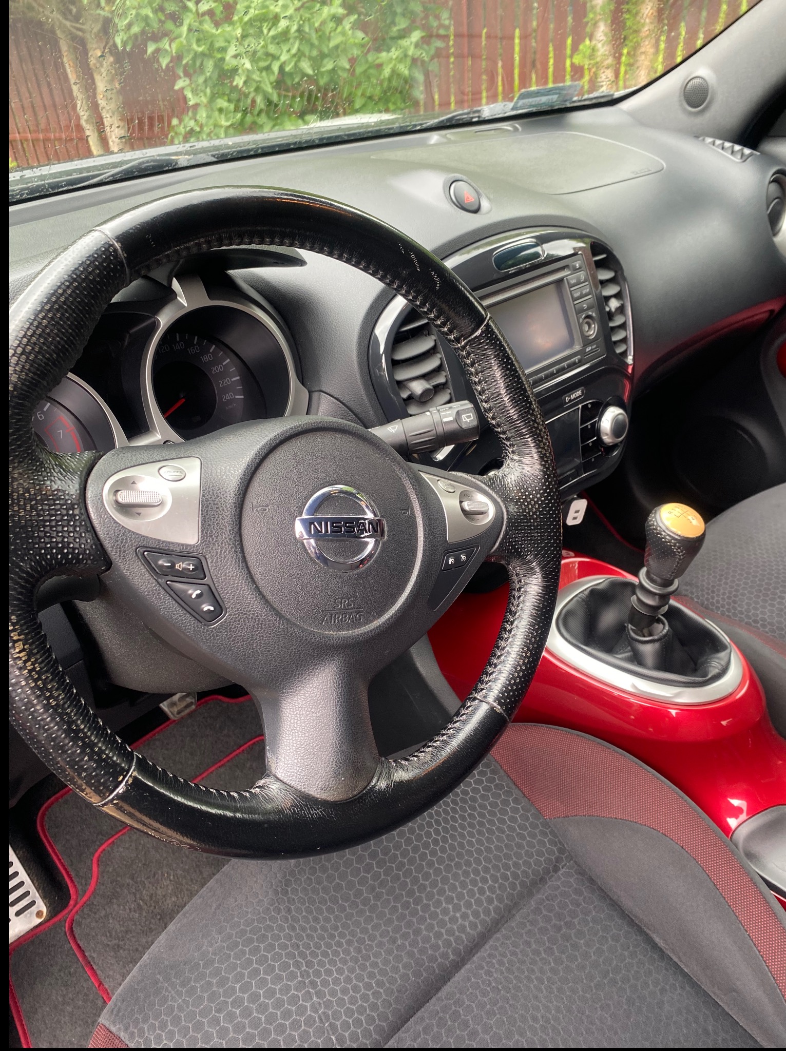 Wnętrze Nissana Juke: kierownica z perforowanej skóry, deska rozdzielcza z ekranem, fotel z szarej, heksagonalnej tkaniny i czerwone akcenty. Widok z perspektywy kierowcy.