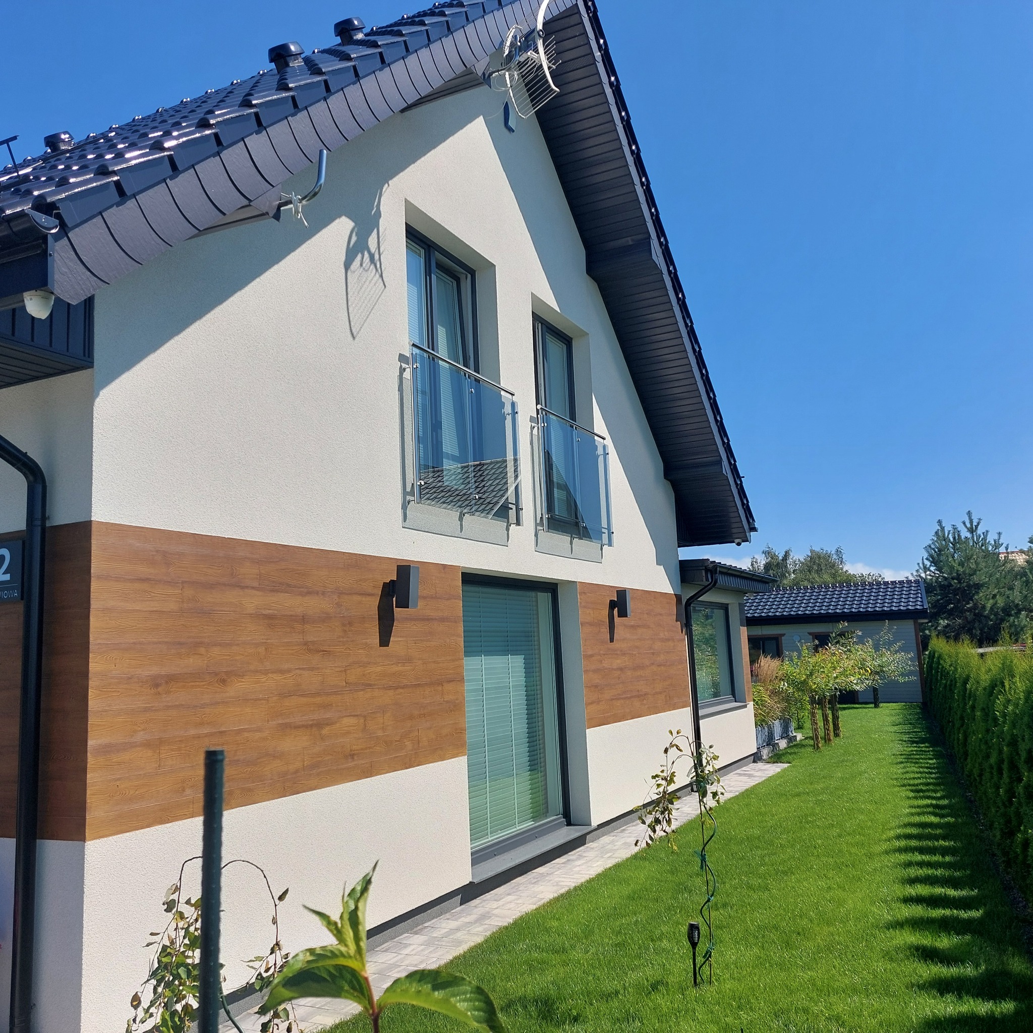 Nowoczesny dom z drewnopodobną elewacją, balkonami ze szkła i równo przystrzyżonym trawnikiem w słoneczny dzień. Widoczna antena satelitarna i oświetlenie zewnętrzne.