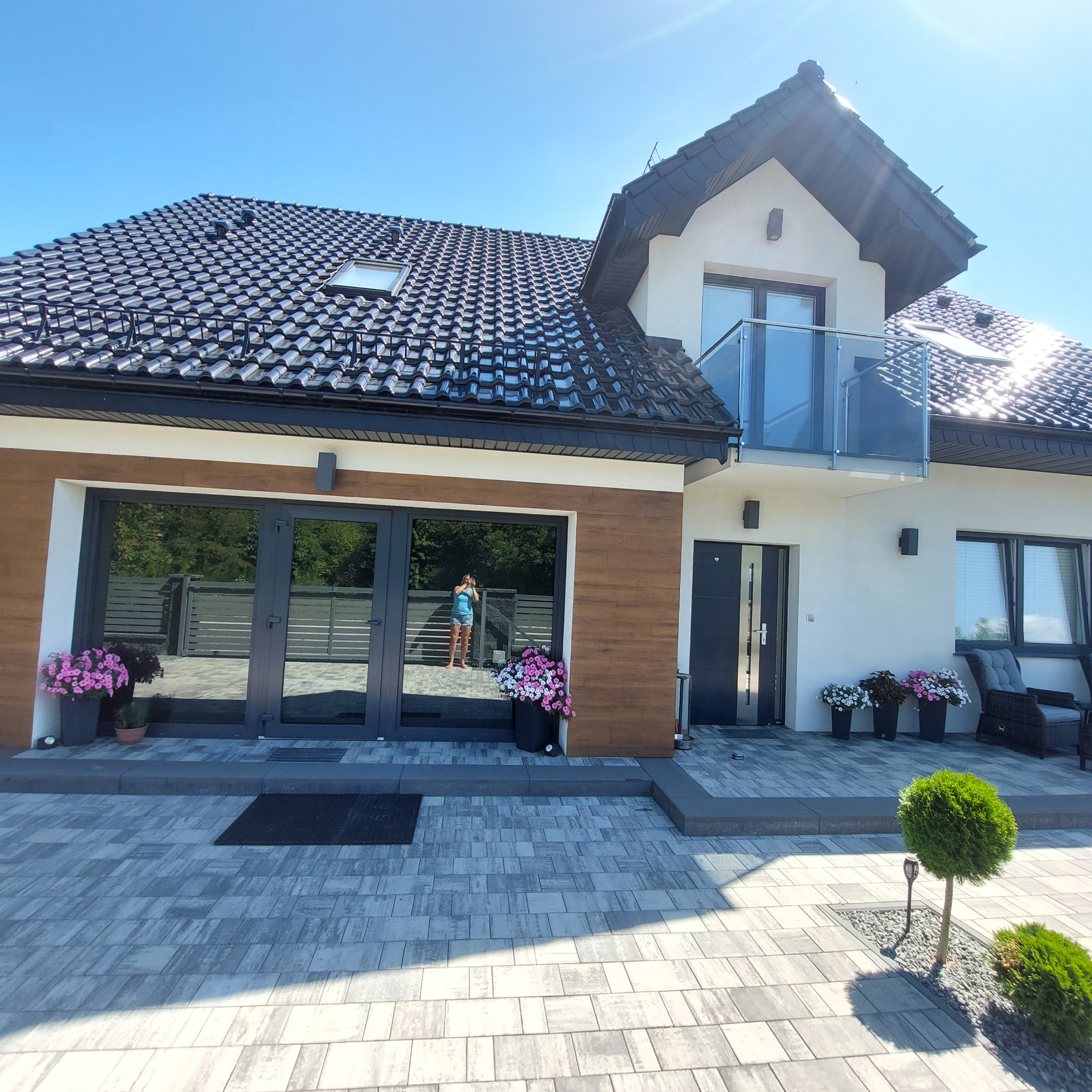 Nowoczesny dom z balkonem i dużymi oknami, otoczony kostką brukową. Widoczna elewacja w bieli i drewnie, z kwiatami w donicach. W oknie odbija się osoba robiąca zdjęcie.