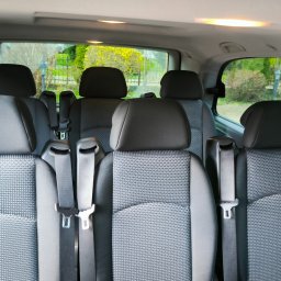 GROM - Wnętrze przestronnego busa z rzędami szarych foteli z zagłówkami i pasami bezpieczeństwa, widok z tyłu pojazdu na zieleń za oknami.