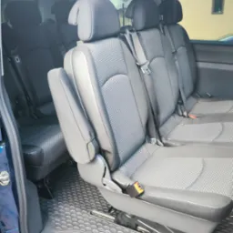 Wnętrze przestronnego busa z trzema rzędami szarych foteli z zagłówkami i pasami bezpieczeństwa, widok z otwartych drzwi bocznych.