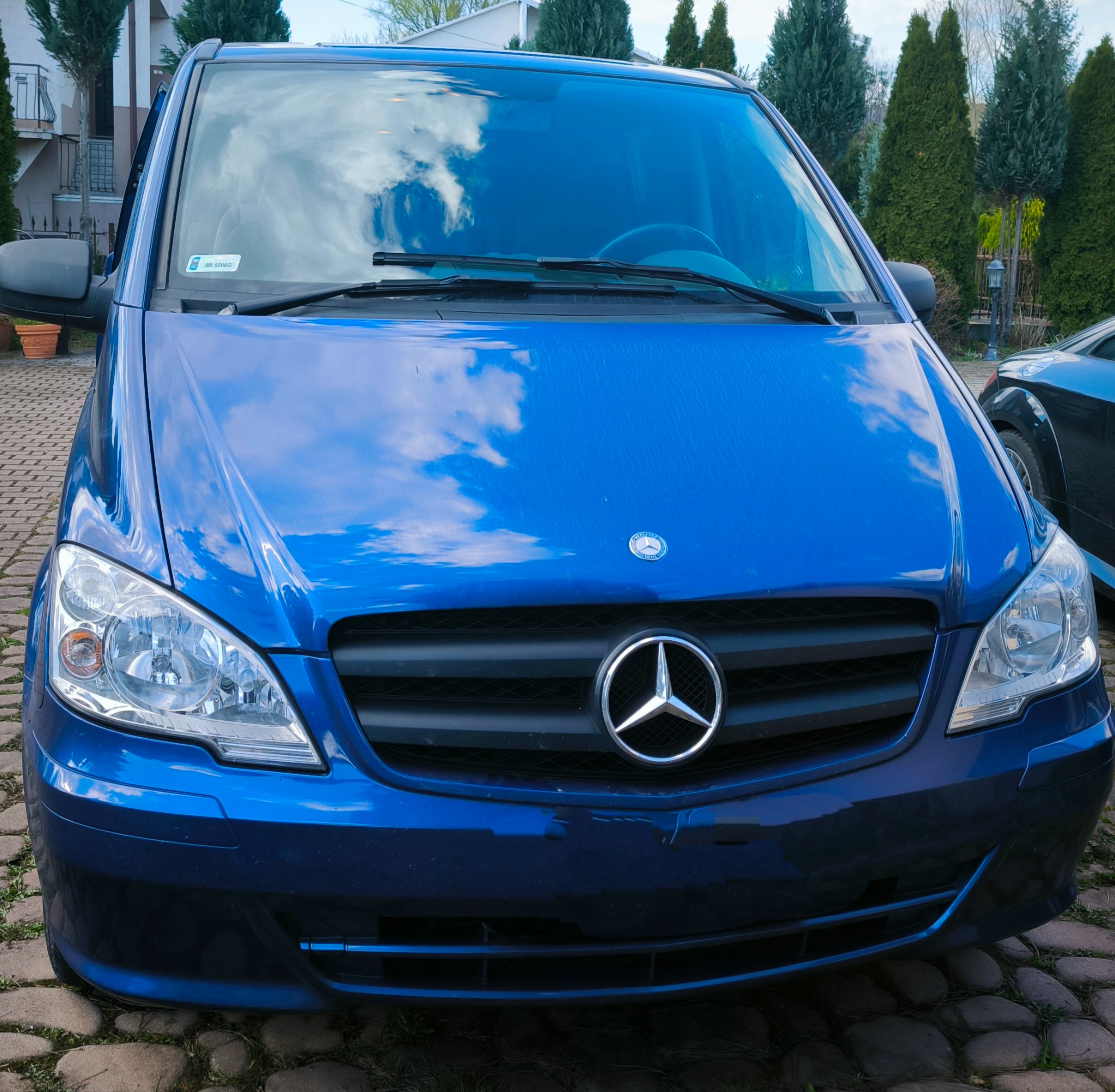 Niebieski Mercedes Vito z widocznym odbiciem chmur na przedniej szybie, zaparkowany na brukowanej nawierzchni, z zielenią w tle.