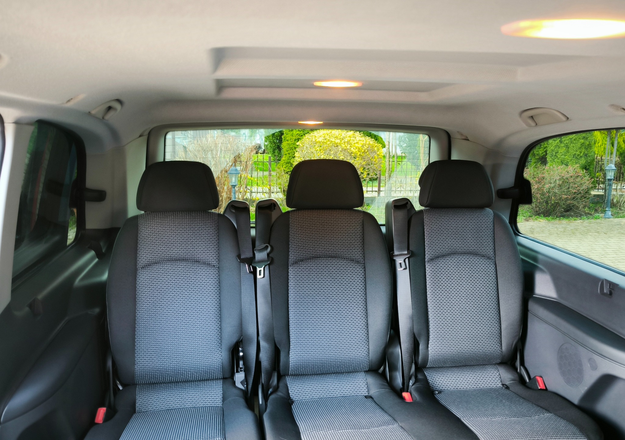 Wnętrze busa z trzema rzędami foteli w szaro-czarnej tapicerce. Widok z tyłu pojazdu na siedzenia i okno z zielenią w tle. Oświetlenie sufitowe.