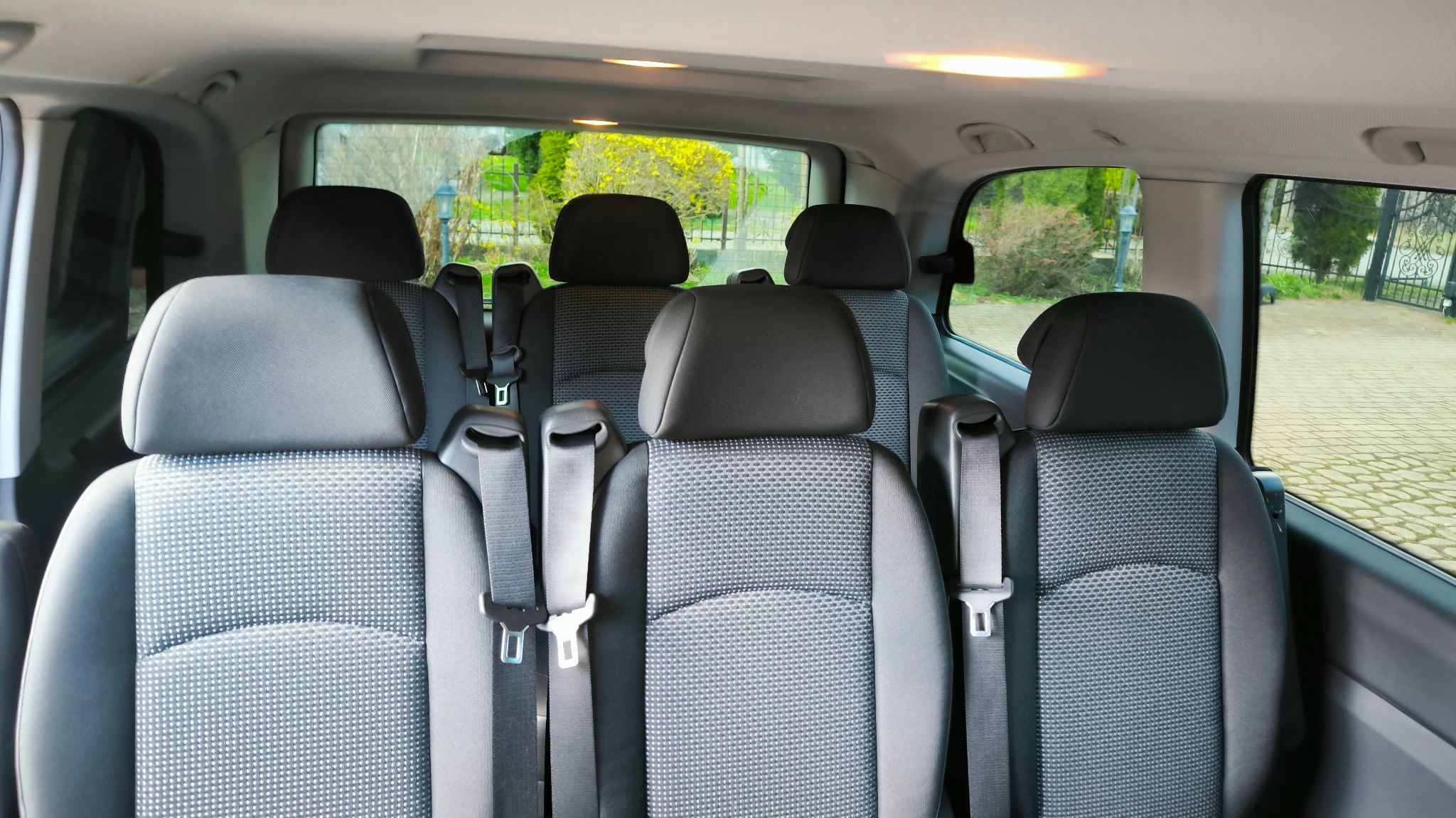 Wnętrze przestronnego busa z rzędami szarych foteli z zagłówkami i pasami bezpieczeństwa, widok z tyłu pojazdu na zieleń za oknami.