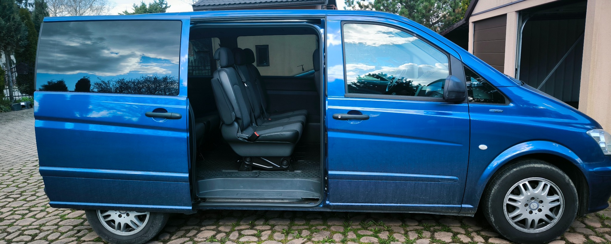 Niebieski bus Mercedes Vito z otwartymi drzwiami bocznymi, prezentujący wnętrze z fotelami pasażerskimi. Widoczne otoczenie zewnętrzne, kostka brukowa i garaż.