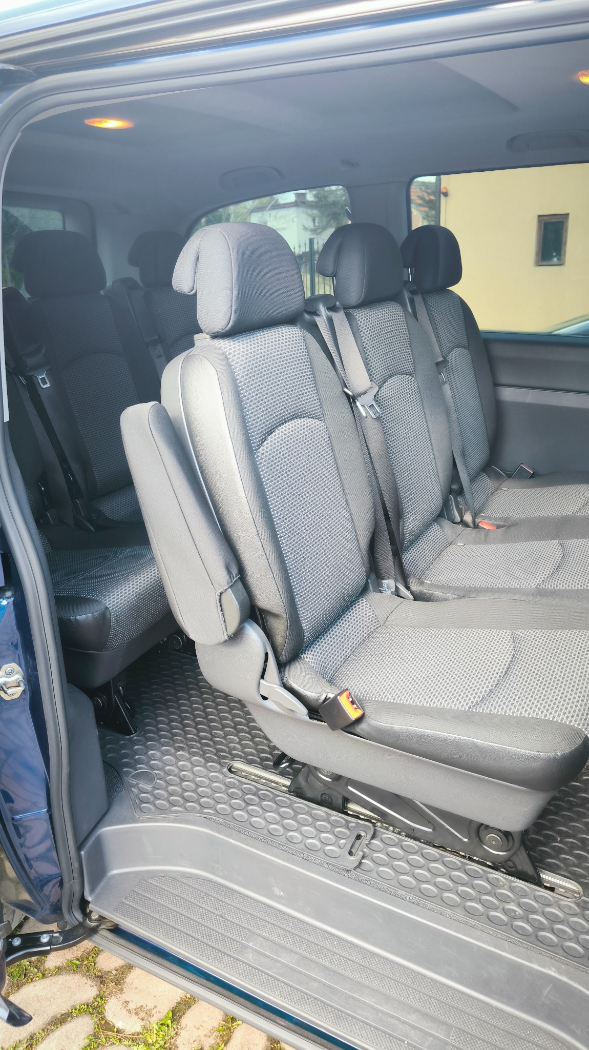 Wnętrze przestronnego busa z trzema rzędami szarych foteli z zagłówkami i pasami bezpieczeństwa, widok z otwartych drzwi bocznych.