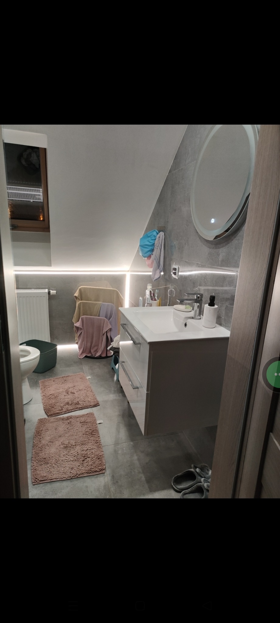 Nowoczesna łazienka na poddaszu w Ostrowi Mazowieckiej: szare płytki, biała umywalka z szafką i okrągłe lustro z podświetleniem LED. Uwagę przykuwają dwa puszyste dywaniki.