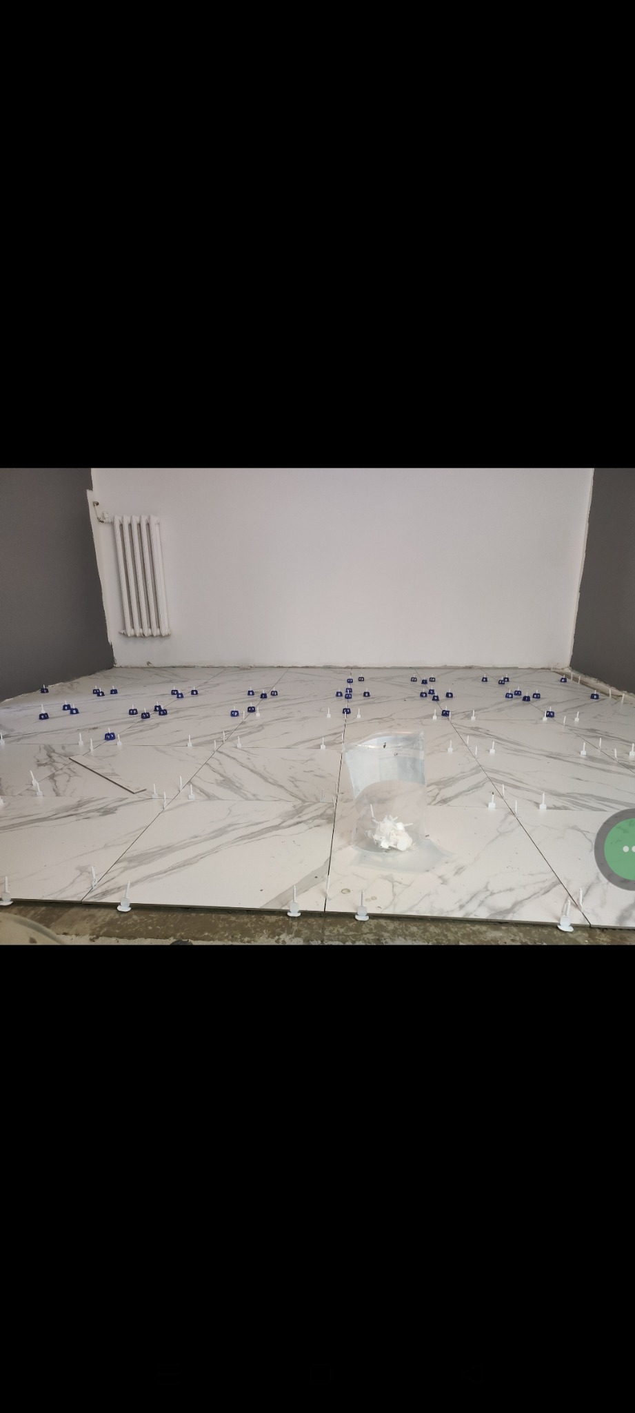 Układanie płytek podłogowych imitujących marmur, z użyciem klipsów i klinów poziomujących. Widoczny fragment ściany z grzejnikiem.