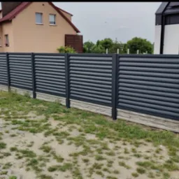 Nowoczesne ogrodzenie metalowe, poziome lamele w kolorze grafitowym, podmurówka z imitacji kamienia. Widok na domy w tle, trawnik z rzadką trawą.