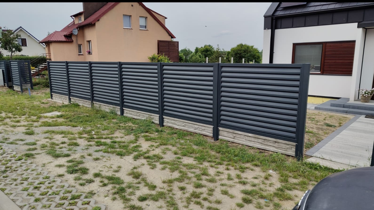 Nowoczesne ogrodzenie metalowe, poziome lamele w kolorze grafitowym, podmurówka z imitacji kamienia. Widok na domy w tle, trawnik z rzadką trawą.