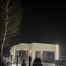 Nowoczesny dom z płaskim dachem oświetlony nocą, widoczny zza metalowego ogrodzenia. Minimalistyczna architektura z podświetleniem elewacji i ścieżki.