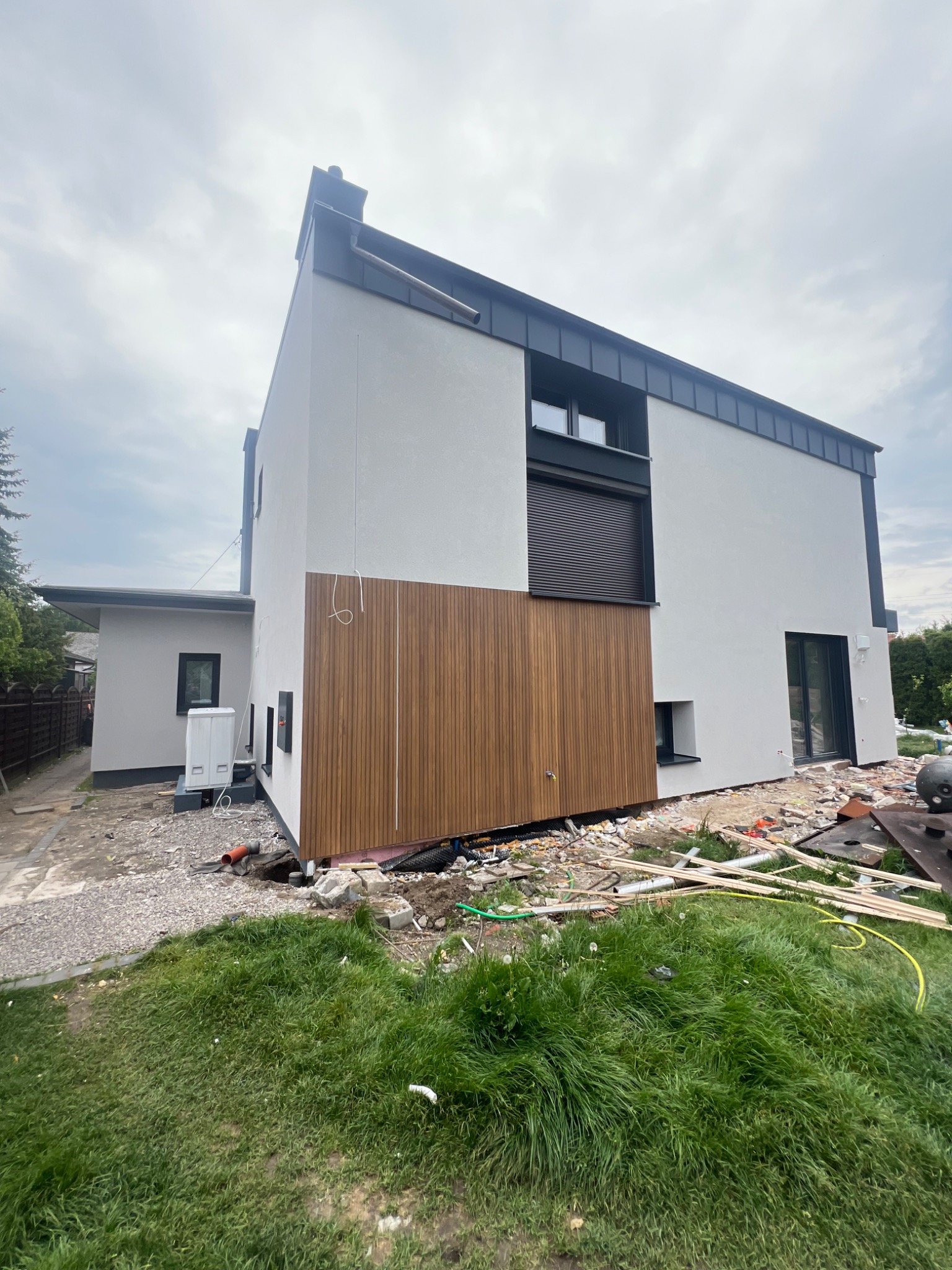 Nowoczesna elewacja domu w trakcie wykończenia: biały tynk kontrastuje z drewnianą okładziną. Widoczne narzędzia i materiały budowlane na trawniku.
