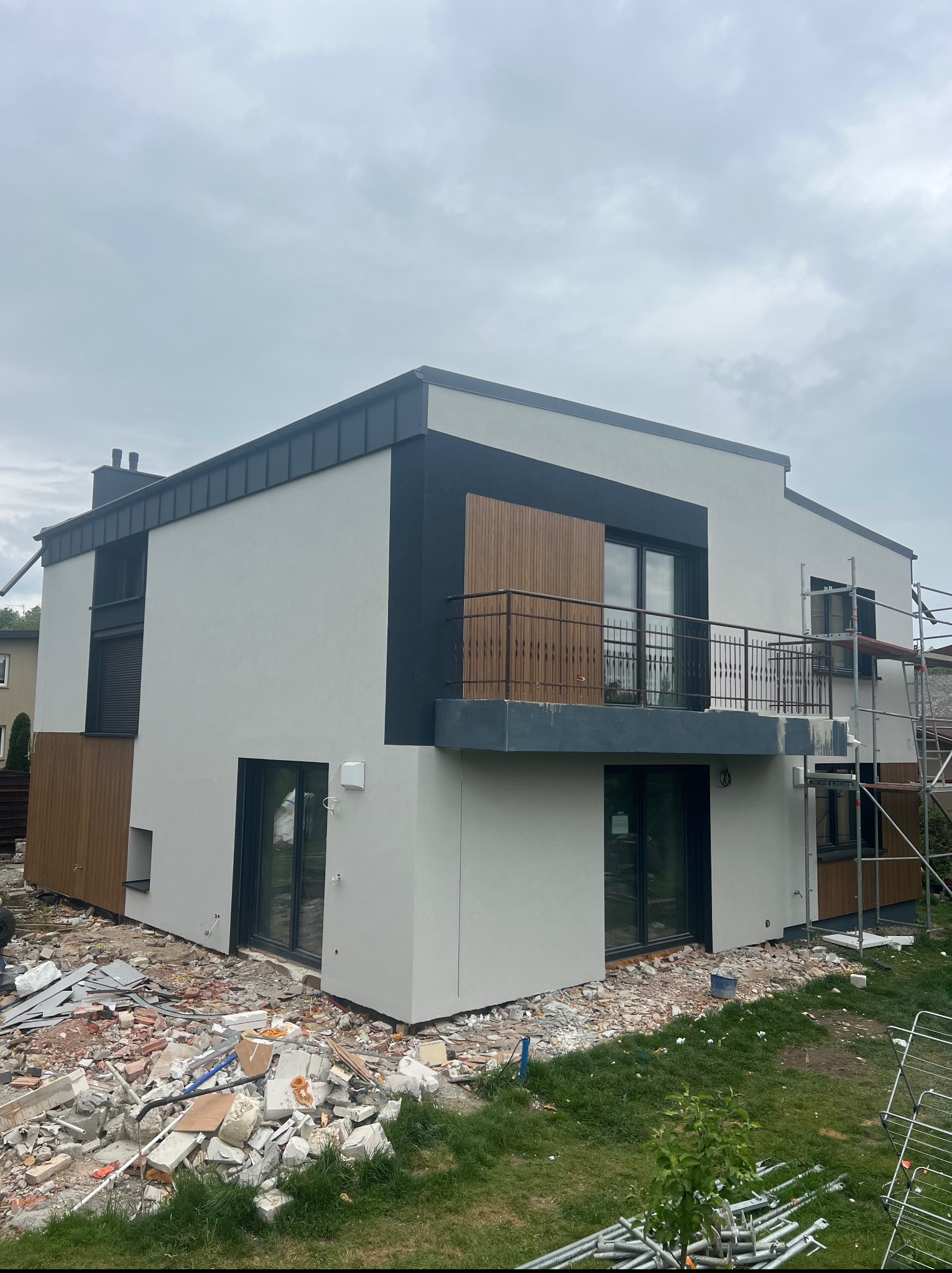 Nowoczesny dom z wykończoną elewacją w odcieniach szarości i brązu, z balkonem i rusztowaniem, otoczony gruzem po pracach budowlanych.