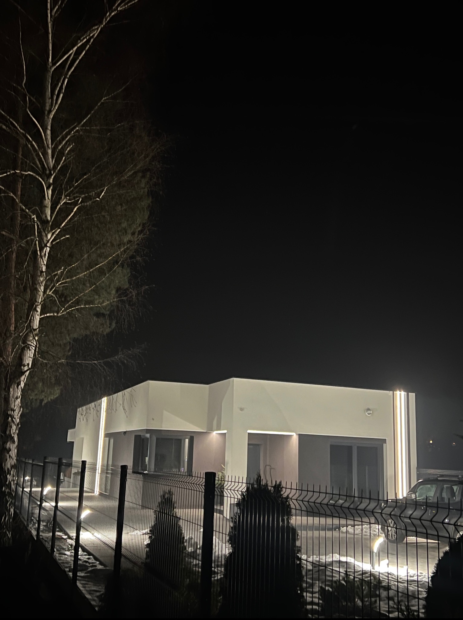 Nowoczesny dom z płaskim dachem oświetlony nocą, widoczny zza metalowego ogrodzenia. Minimalistyczna architektura z podświetleniem elewacji i ścieżki.