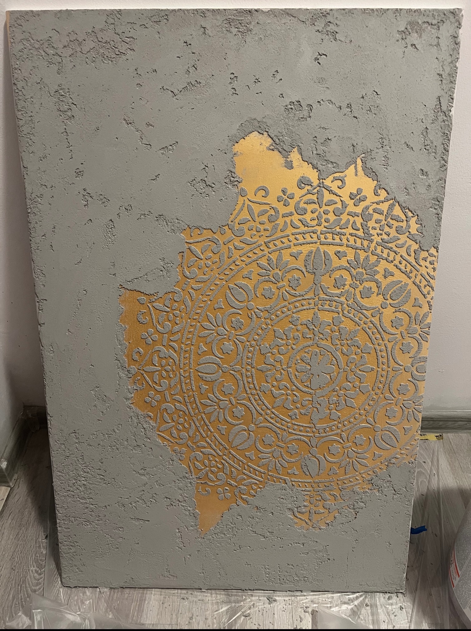 Dekoracyjna ściana z szarą, teksturowaną powierzchnią i odsłoniętym złotym ornamentem w stylu mandali. Fragment podłogi zabezpieczony folią malarską.