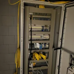 Otwarta szafa sterownicza z licznymi przewodami i elementami elektronicznymi. Żółte kable zwisają po lewej stronie, kontrastując z szarą ścianą z cegieł.