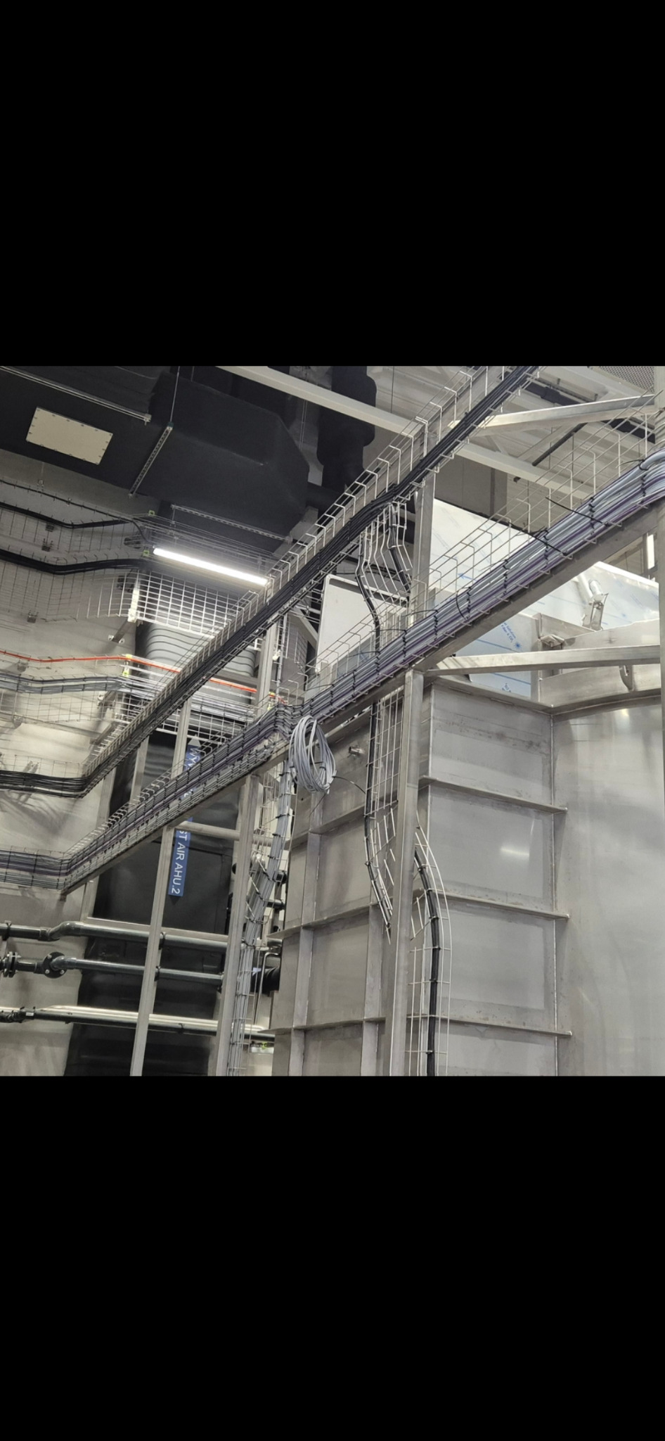 Rozbudowana instalacja elektryczna z kablami prowadzonymi w korytach i po drabinkach w hali przemysłowej. Widok z dołu na skomplikowany system.