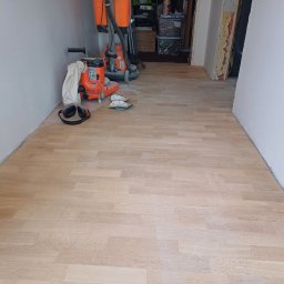 Andrzej Zielski - Jasna, odnowiona podłoga parkietowa po cyklinowaniu, z widocznymi maszynami i akcesoriami do obróbki drewna w tle. Widoczne ślady pyłu po szlifowaniu.