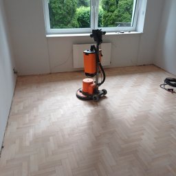 Andrzej Zielski - Parkiet ułożony w jodełkę po obróbce. Na środku pomieszczenia stoi pomarańczowa maszyna do obróbki parkietu. Widok na zieleń za oknem.