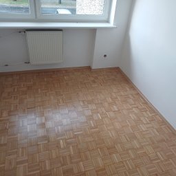Andrzej Zielski - Parkiet w jodełkę po cyklinowaniu w pustym, jasnym pokoju z grzejnikiem pod oknem. Widoczne gładkie, drewniane klepki i świeżo pomalowane ściany.