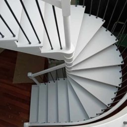 Białe, spiralne schody z czarnymi balustradami, widok z góry. Drewniana podłoga w tle. Minimalistyczny design, świeżo po renowacji. Jasne oświetlenie.
