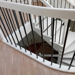 Białe, odnowione schody spiralne z czarnymi balustradami, widok z góry na drewnianą podłogę. Świeżo pomalowane, z widocznymi detalami wykończenia.