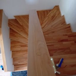 Spiralne schody z jasnego drewna po cyklinowaniu, widok z góry. Na dole widoczne narzędzia i zabezpieczenia podłogi. Charakterystyczny wzór ułożenia desek.