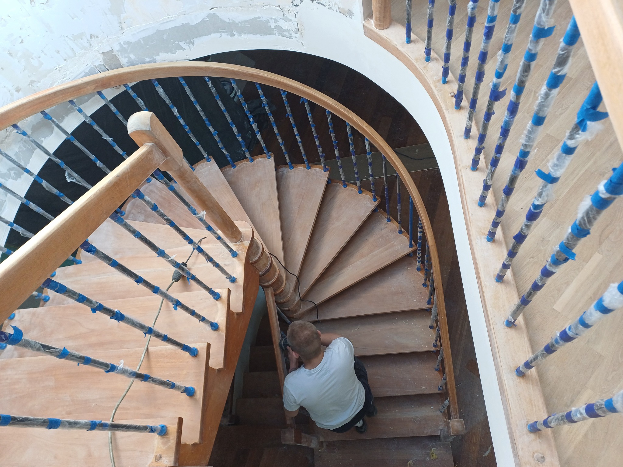 Prace renowacyjne na drewnianych, kręconych schodach. Widok z góry na pracownika szlifującego stopnie. Balustrady zabezpieczone taśmą malarską.