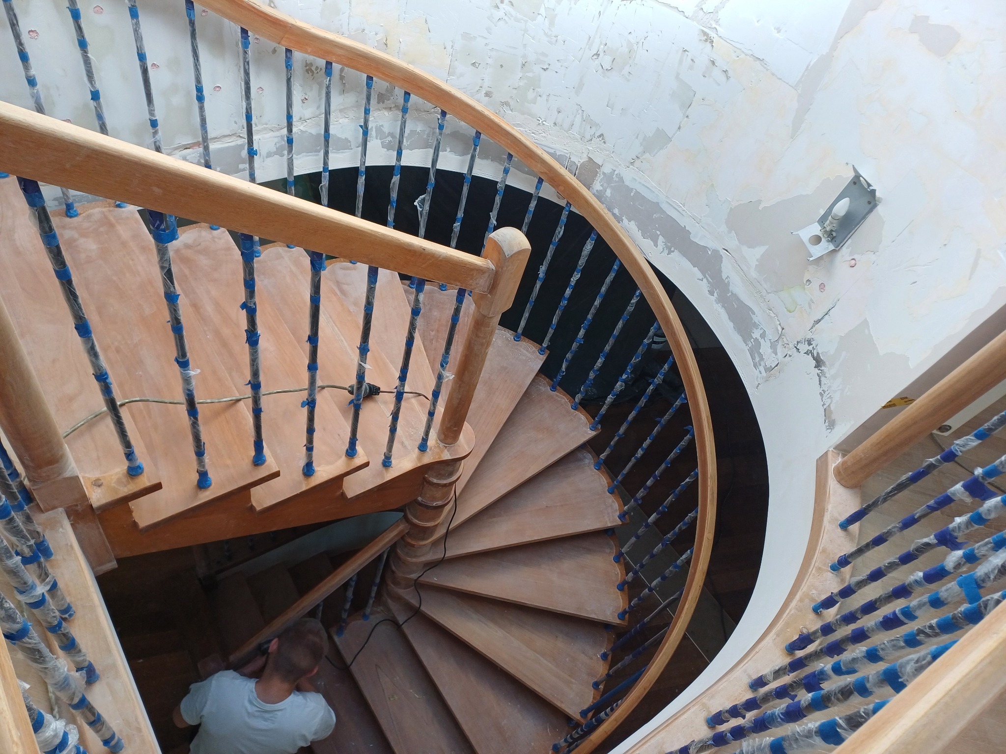 Drewniane schody spiralne w trakcie renowacji, zabezpieczone barierki, widoczny fragment pracownika, surowe ściany w tle. Ujęcie z góry.