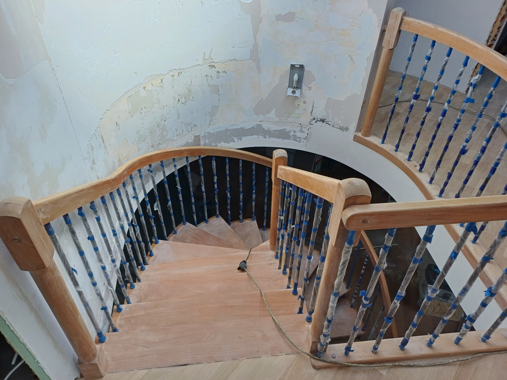 Spiralne schody drewniane po renowacji, zabezpieczone folią i taśmą malarską. Widok z góry na odnowione stopnie i balustrady. Ściana w tle przygotowana do malowania.