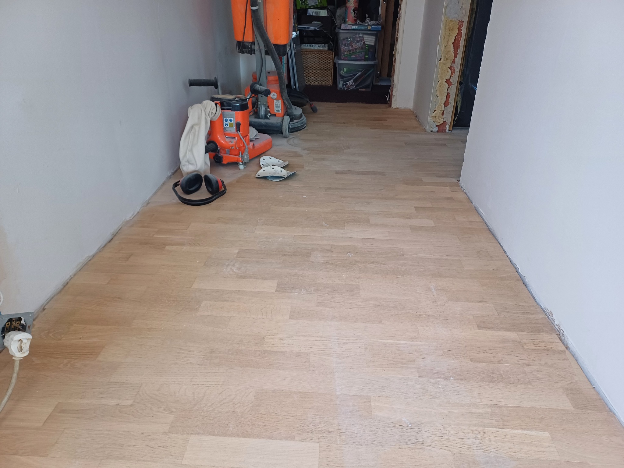 Jasna, odnowiona podłoga parkietowa po cyklinowaniu, z widocznymi maszynami i akcesoriami do obróbki drewna w tle. Widoczne ślady pyłu po szlifowaniu.
