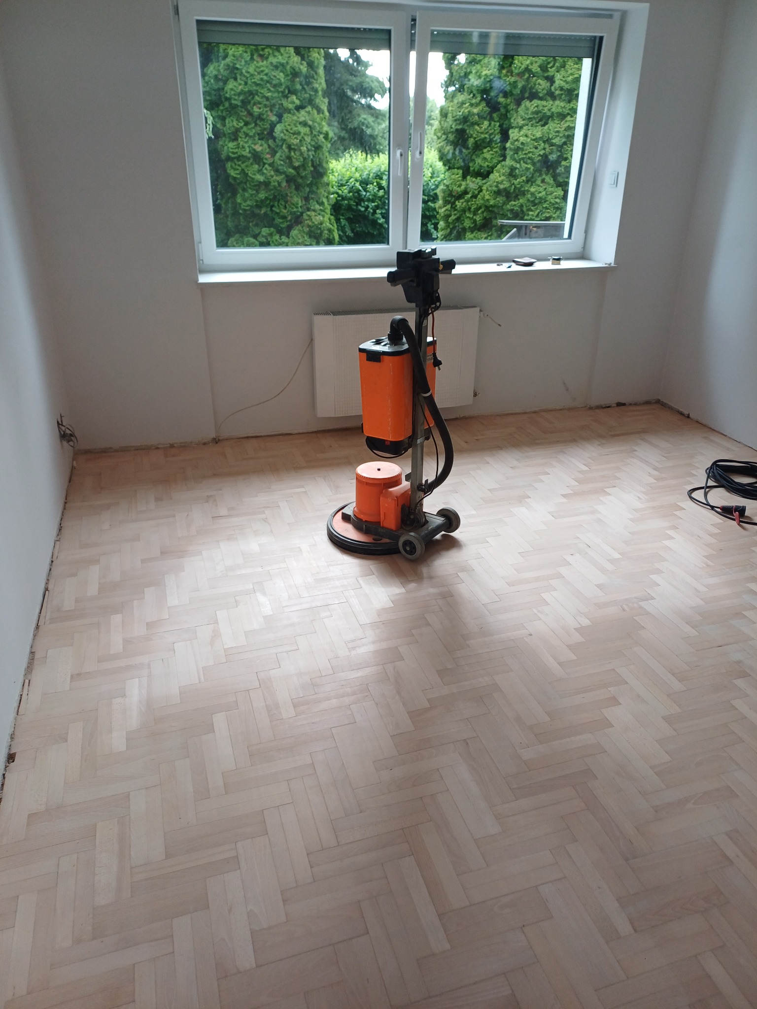 Parkiet ułożony w jodełkę po obróbce. Na środku pomieszczenia stoi pomarańczowa maszyna do obróbki parkietu. Widok na zieleń za oknem.